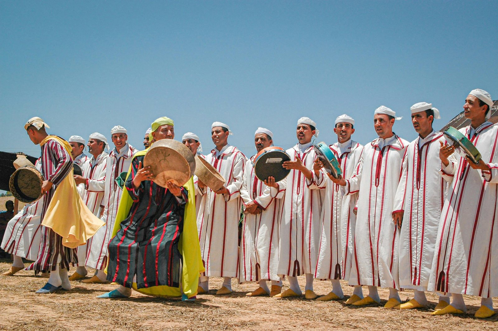 A group of Moroccan musicians in traditional attire performing outdoors with percussion instruments under a clear sky.